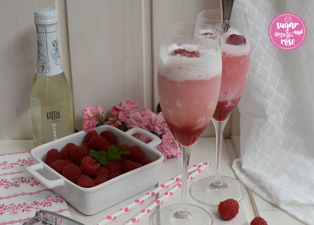 Zwei Sektgläser mit schäumendem Roseneis-Float, eine kleine Kattus-Sektflasche dahinter, davor eine weiße, quadratische Keramikschale mit Himbeeren, ein rosa geblümtes Geschirrtuch und zwei Papierstrohhalme mit rosa Herzerl drauf