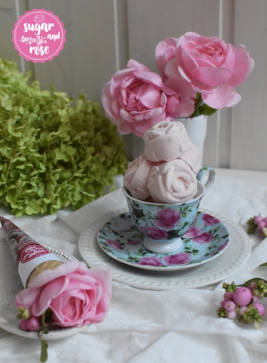 Roseneis in Form von kleinen Rosenblüten aufgetürmt in einer hellblauen Mokkatasse mit Untertasse, beides mit Rosenmotiven, dahinter eine weiße Vase mit zwei rosa Rosenblüten, davor ein Waffelhörnchen mit einer Rosenblüte