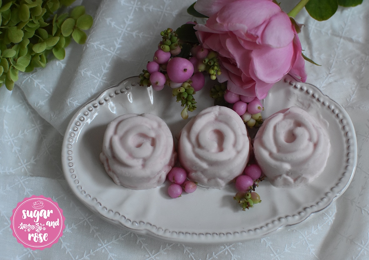 Drei Stück Roseneis in Rosen-Pralinenform auf einem ovalen weißem Keramikteller im Vintage-Style, daneben Rosenblüten und rosa Kugeln vom Liebesperlenstrauch