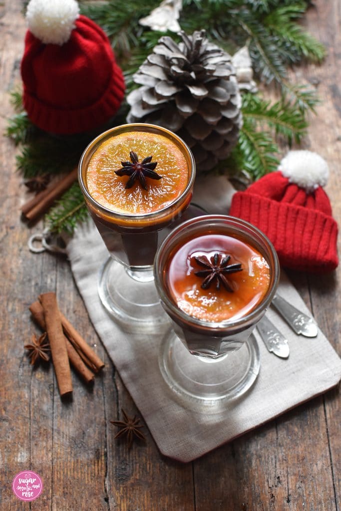 Zwei alte Teegläser aus Pressglas mit Stiel, gefüllt mit Punsch, Orangenscheibe und Sternanis, daneben auf einem rustikalen Brett Tannenzweige, ein Zapfen, zwei kleine rote Wollmützen mit weißer Quaste, davor Zimtstangen