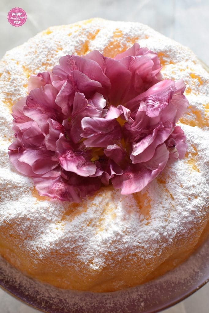 Detailansicht des Topfengugelhupfs, von oben fotografiert, mit einer dicken Puderzuckerschicht und einer lila Tulpenblüte in der Gugelhupfmitte als Dekoration