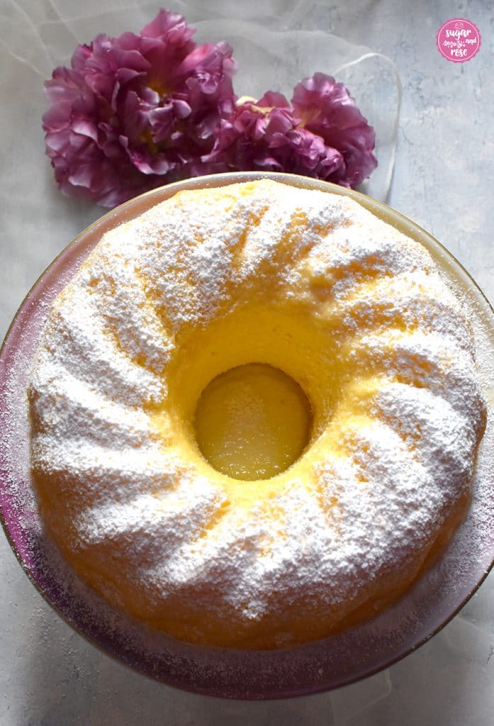 Sonnig gelber Topfengugehlhupf auf rosa Tortenständer, daneben lila Tulpen. Der Gugelhupf leuchtet von der Mitte heraus sonnengelb und wirkt ursaftig.