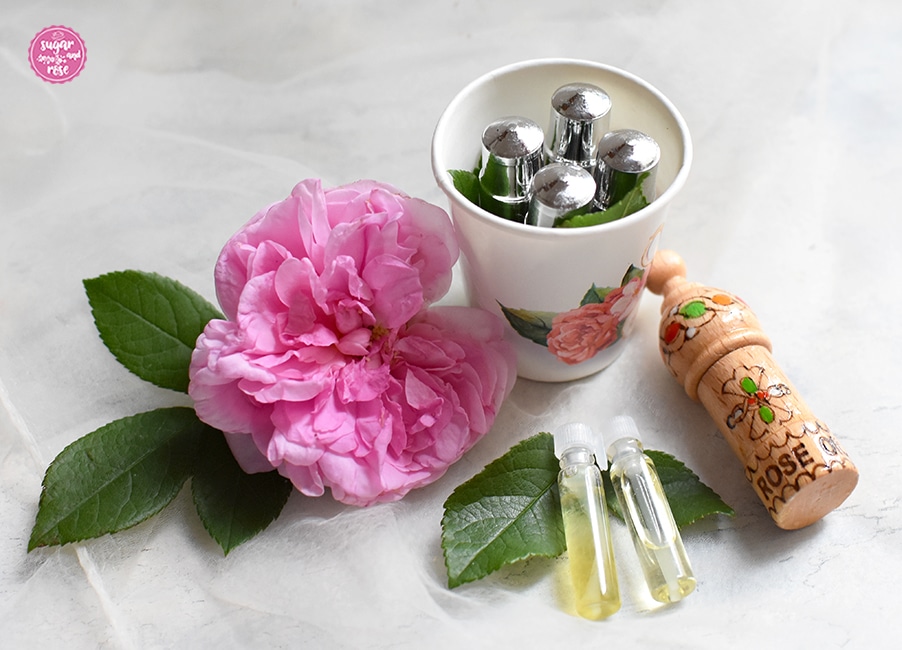Zweierlei Rosenöl in kleine Philolen, zwei im Vordergrund durchsichtig, vier in metallenen Behältern und eine in einem Holzbehälter mit der Aufschrift Rose Oil Bulgaria, dazu eine große Rosenblüte der historischen Rose Ispahan