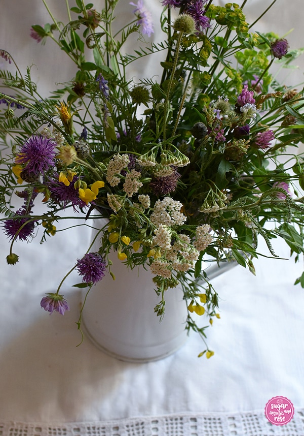 Wiesenblumenstrauß mit gelbem Hornklee, violetten Flockenblumen, rotem Hornklee und fliederfarbenen Acker-Weitwenblumen und weißer Schafgarbe in einer weißen Vintagekanne mit Henkel auf einem alten weißen Handtuch mit gehäckeltem weißem Rand