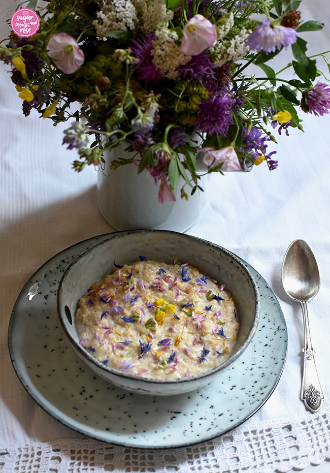 Sommer-Porridge mit bunten Wiesenblumen in einer einer grauen Schale mit Silberlöffel, dahinter ein Wiesenblumenstrauß mit gelbem Hornklee, violetten Flockenblumen und weißer Schafgarbe in einer grauen Vase
