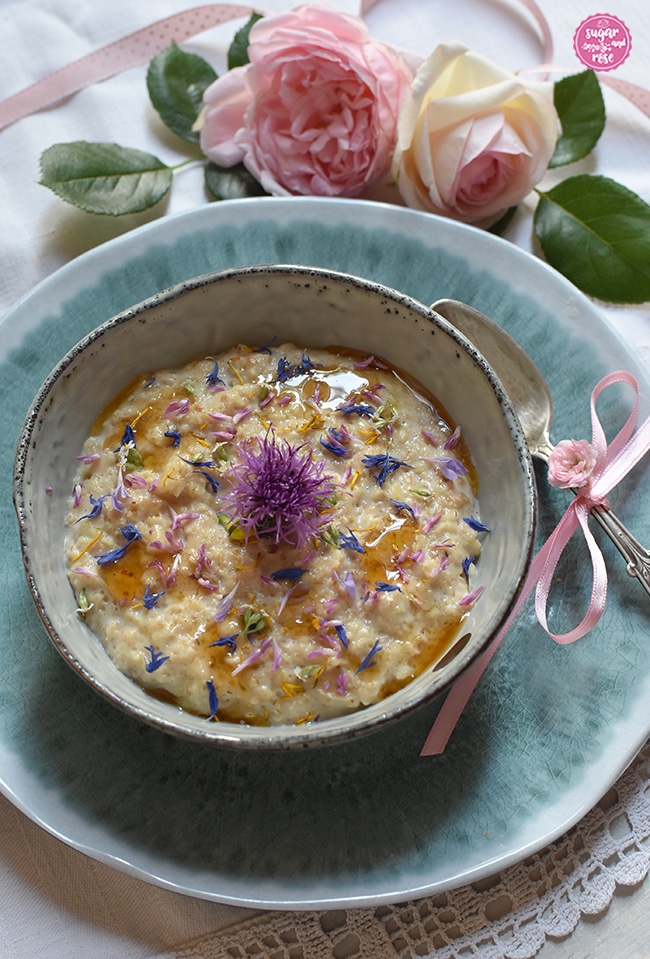 Sommer-Porridge mit bunten Wiesenblumen in einer einer grauen Schale mit Silberlöffel, dahinter zwei rosa Rosen mit einem rosa getupften Bändchen; die Schale steht auf einem türkis marmorierten Teller. Das Porridge ist garniert mit blauen Kornblumenblüten, rotem Hornklee und gelbem Löwenzahn, darüber etwas Löwenzahnsirup geträufelt