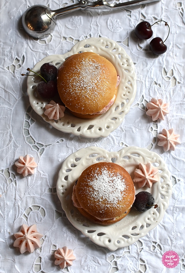Zwei Eisburger (Brioche con Gelato) auf jeweils einem herzförmigen Keramikteller mit Lochmusterdekor auf einem weißen Platzset mit Lochstickerei, dekoriert mit Kirschen und rosa Baisertuffs, dahinter als Deko ein leerer Eisportionierer aus Stahl