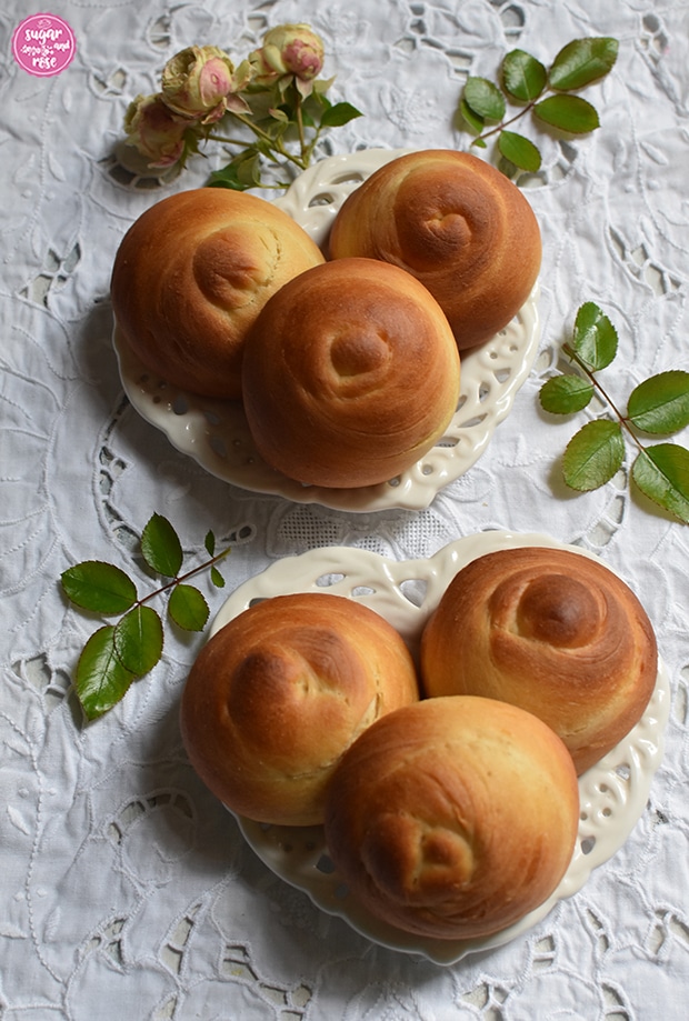 Zwei herzförmige Keramikteller mit jeweils drei frisch gebackenen Briocheknöpfen, stehend auf einem weißen Platzset mit Lochstickerei, dekoriert mit einigen Rosenblättern und einem kleinen Rosensträußchen in grün-rosa