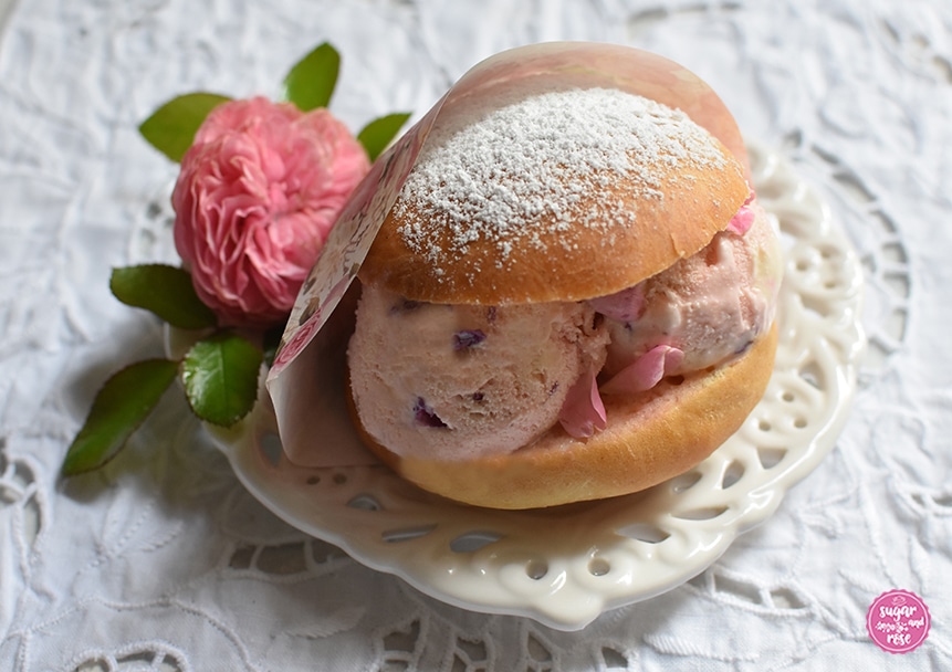 Eisburger (Brioche con Gelato) in einer Papiertüte mit Rosendekor, liegend auf einem Keramikteller auf einem einem weißen Platzset mit Lochmuster, dekoriert mit einer rosa Rosenblüte der Rose Leonardo de Vinci