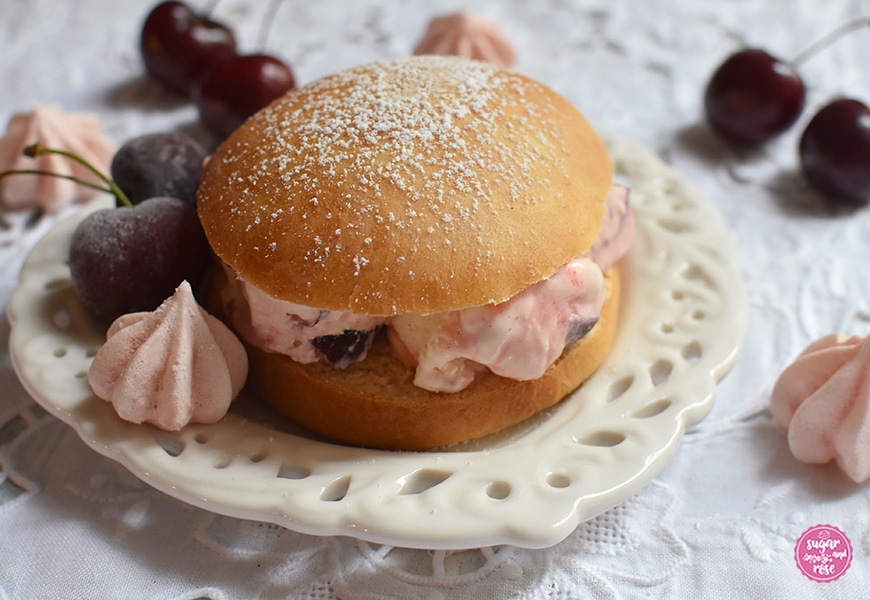 Ein mit Puderzucker bestäubter Eisburger (Brioche con Gelato) auf einem herzförmigen Keramikteller, gefüllt mit rosa-weiß marmoriertem Kirscheneis, dekoriert mit Kirschen und rosa Baisertuffs