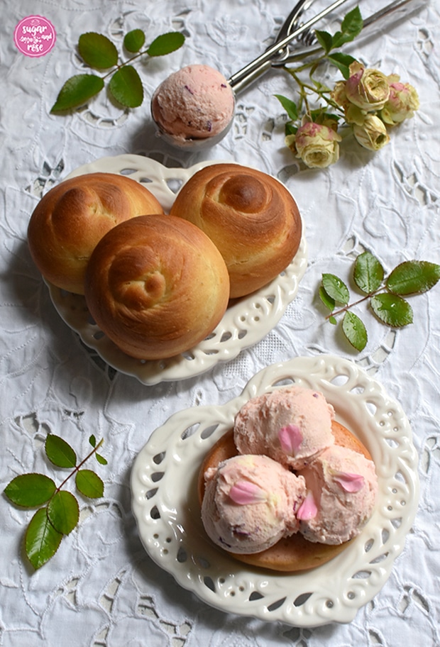 Drei rosa Roseneiskugeln auf einem aufgeschnittenen Brioche, liegend auf einem herzförmigen weißen Vintage-Keramikteller, dahinter ein weiterer Keramikteller mit drei ungefüllte Briocheweckerln von oben und ein Eisportionierer mit einer Kugel Roseneis. Als Deko ein Zweig mit grün-rosa Röschen der Rose Lovely Green