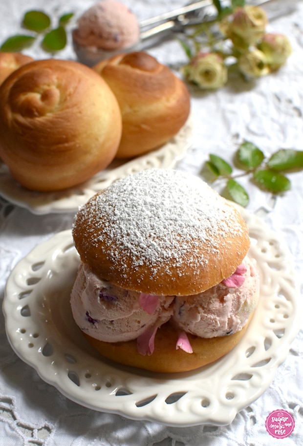 Eisburger (Brioche con Gelato) in gefüllt mit Roseneiskugeln auf Vintageteller, dahinter ein ebensolcher Keramikteller mit ungefüllten Brioschknöpfen, ein Eisportionierer mit einer Kugel Eis und daneben kleine grüne Rosen