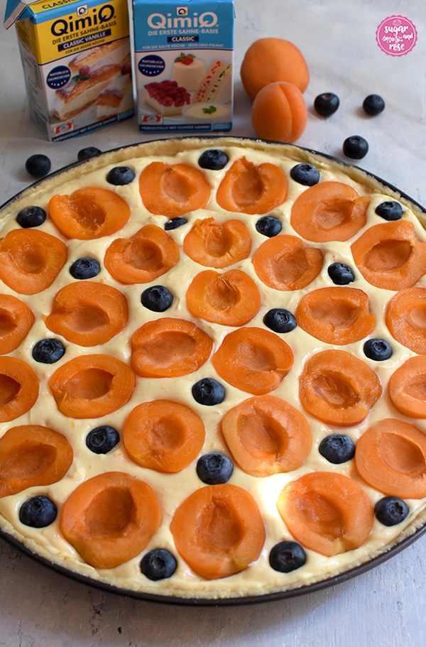 Runde Tarteform mit Marillen-Tarte vor dem Backen. Halbierte Marillen und Heidelbeeren ruhen im Topfenbeet. Hinter der Tarteform stehen zwei Packungen Qimiq