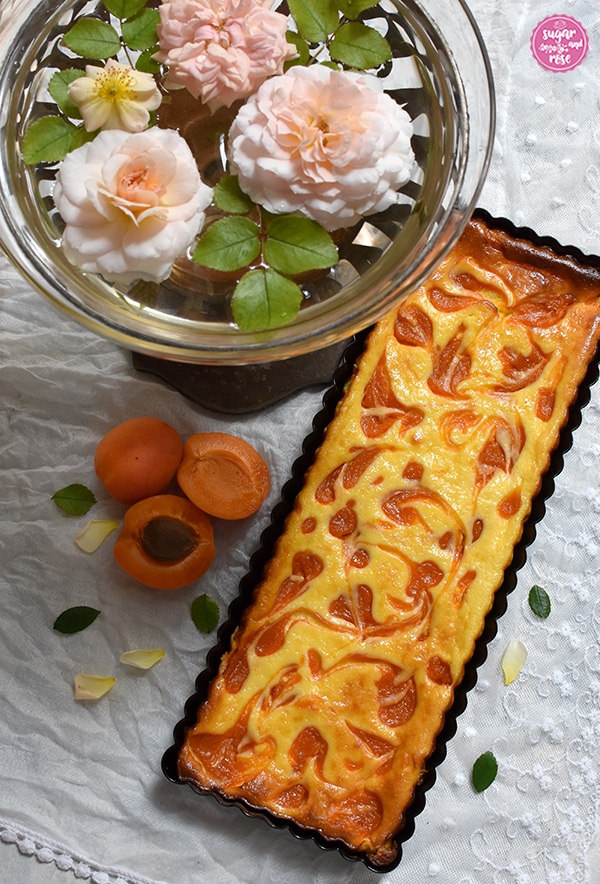 Marillen-Tarte in länglicher Tarteform auf weißem Spitzenschale, daneben eine Jugendstilschale mit Glaseinsatz und schwimmenden apricotfarbenen Rosenköpfen