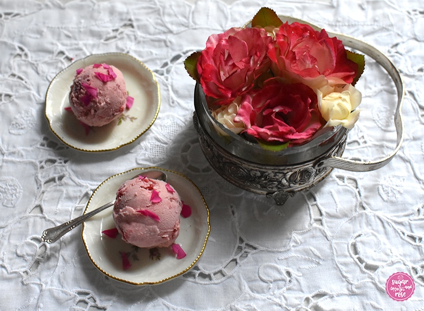 Zwei rosa Roseneiskugeln auf zwei winzig kleinen Keramiktellern mit Goldrand, dazu ein kleiner Vintagesilberlöffel. Daneben eine Jugendstilschale mit Glaseinsatz und Henkel, gefüllt mit drei rot-weißen Rosenblüten der Rose Blumenstadt Tulln und einigen kleinen cremefarbenen Rosenblüten