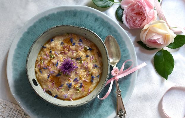 Porridge mit bunten Wiesenblumen in einer Steingut-Bowl auf türkisem Teller, daneben ein alte Silberlöffel mit rosa Masche und rosa Rosen mit Band im Hintergrund