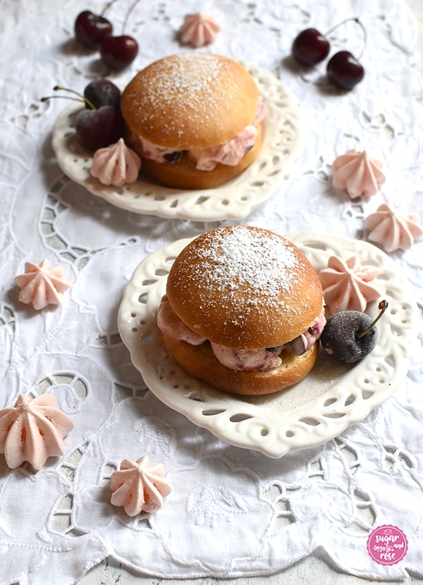 Zwei Eisburger (Brioche con Gelato) auf jeweils einem herzförmigen Keramikteller mit Lochmusterdekor auf einem weißen Platzset mit Lochstickerei, dekoriert mit Kirschen und rosa Baisertuffs
