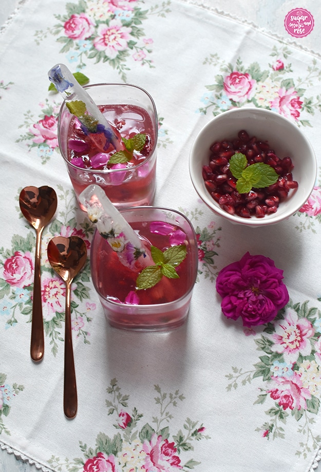 Rosé-Gin-Cocktail mit Granatapfelkernen in zwei eckigen Whiskeygläsern, daneben zwei rosegoldfarbene, herzförmigen Metalllöffelchen, eine Schale mit Granatapfelkernen und Minzblatt, alles auf einer weißen Leinenserviette mit zartrosa Rosenmotiven. In den Cocktailgläsern stecken längliche Eiswürfel mit eingefrorenen Blüten
