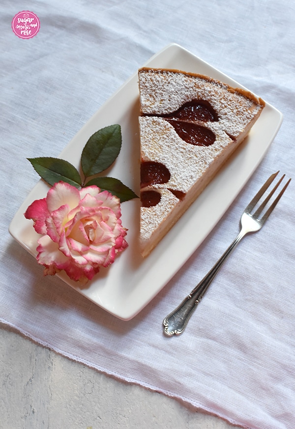 Ein Stück der Herzerl-Ricotta-Tarte mit Zwetschkenmus auf einem eckigen weißen Teller, dekoriert mit einer Rosenblüte (Blumenstadt Tulln, porzellanfarben mit roten Rändern) und grünen Rosenblättern. Daneben eine antike Silber-Dessertgabel auf einer weißen Leinenserviette