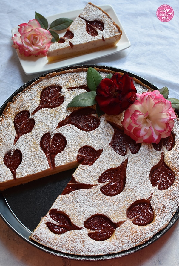 Herzerl-Ricotta-Tarte mit Zwetschkenmus in einer runden Tarteform. Ein Stück fehlt. Es steht im Hintergrund auf einem weißen Keramikteller und man sieht den Anschnitt des Kuchens.