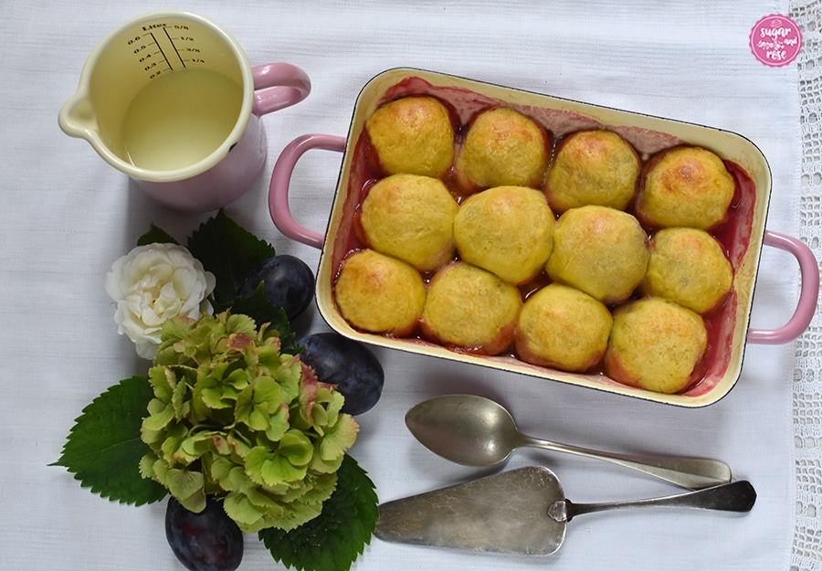 Gebackene Zwetschkenknödel in einer pastellrosa Emailpfanne, von oben fotografiert, neben der rosa Pfanne liegt ein alter Esslöffel und ein alter Tortenheber, daneben ein rosa Emailhäferl mit Maßeinheiten und mit etwas Milch drinnen, vorne eine grüne Hortensienblüte, eine weiße Rose und einige Zwetschken