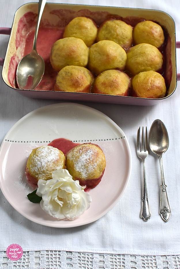 Im Hintergrund eine Pfanne mit gebackenen Zwetschkenknödel, einige Knödel fehlen, in der Pfanne liegt ein alter Esslöffel, danvor ein Dessertteller, zur Hälfte pastellrosa, zur andern Hälfte cremeweiß, darauf zwei Knödel mit Staubzucker, etwas Zwetschkensaft und eine weiße Rosenblüte. Neben dem Teller ein alter Teelöffel und ein Kuchengaberl
