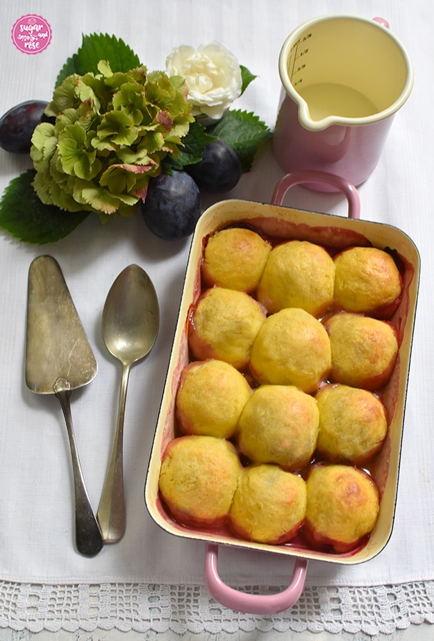 Gebackene Zwetschkenknödel in einer pastellrosa Emailpfanne, daneben ein alter Eßlöffel und Tortenheber, dahinter eine grüne Hortensienblüte, eine weiße Rose, einige Zwetschken und ein rosa Email-Messbecher mit Milch