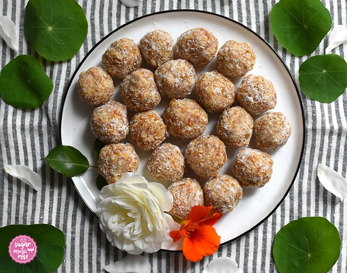 Weißer flacher, runder Teller mit dunkelblauem Rand auf einer grau-weiß gestreiften Leinenserviette. Am Teller einige Marillen-Kokos-Energiekugeln und eine einzelne weiße Rosenblüte sowie eine orange Kapuzinerkresseblüte, rundum grüne Blätter der Kapuinerkresse und einige weiße Rosenpetalen
