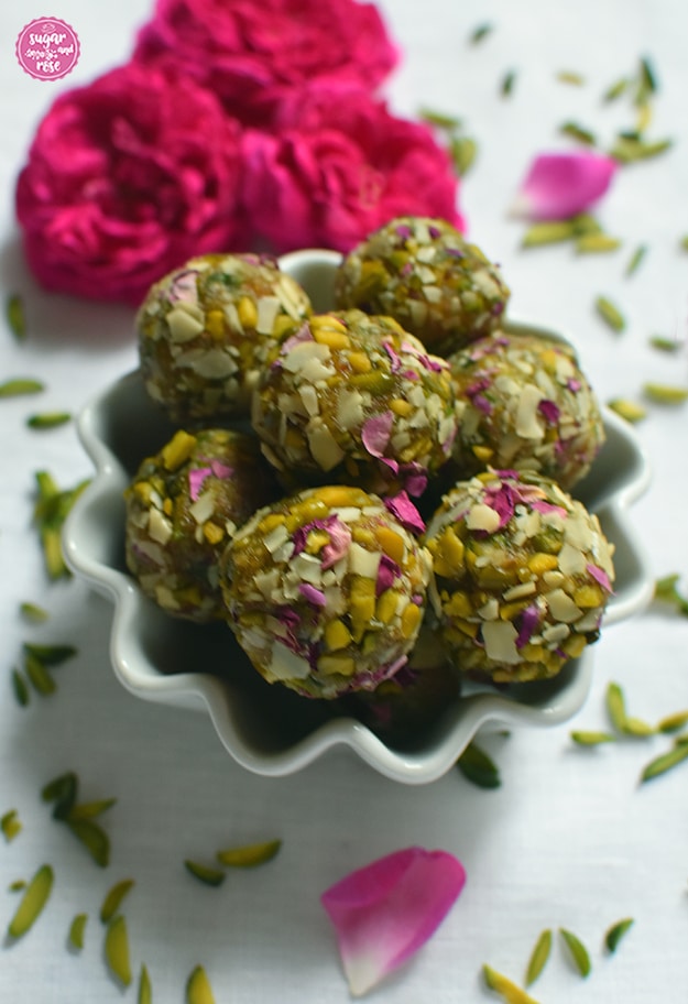 Kleines weißes Porzellanschälchen mit einigen Rosen-Safran-Energiekugeln in Großafufnahme, rundum gehackte grüne Pistazien gestreut, im Hintergrund drei grellpinkfarbene Rosenköpfe