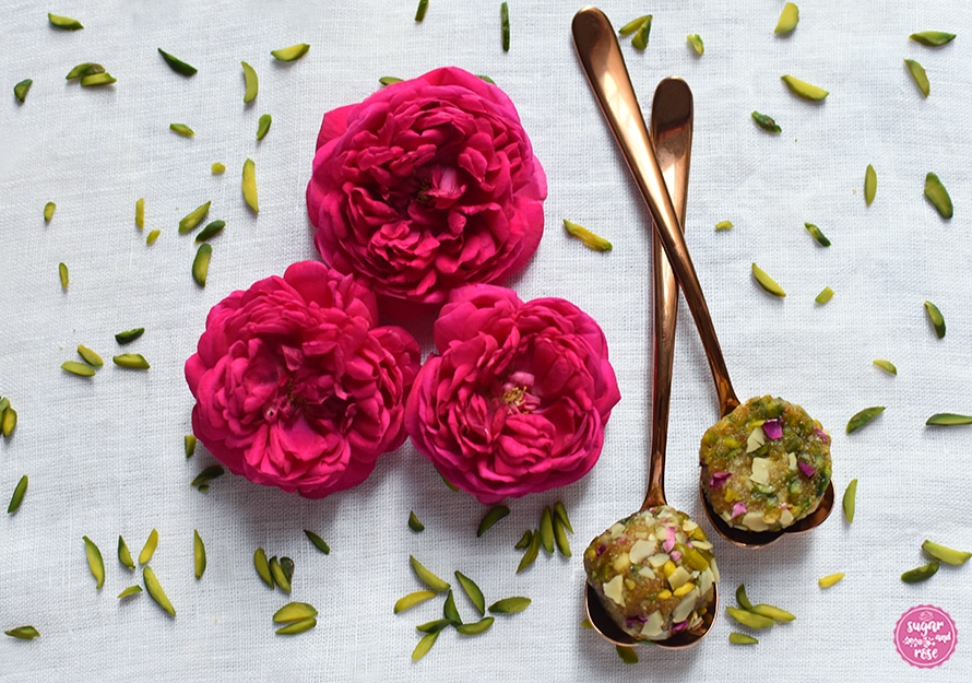 Weißes Leinentischtuch mit grünen Pistazienstiften bestreut, in der Mitte drei pinkfarbene Rosen, daneben zwei kupferfarbene Herzlöffelchen mit überkreuzten Stielen. Auf den Löffeln je ein halbierter Energyball