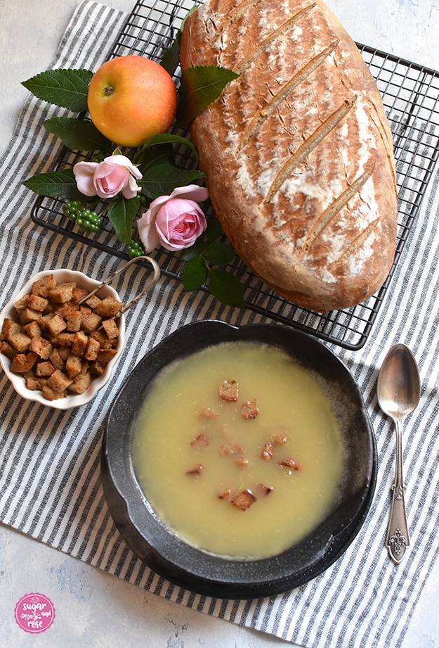 Graue Steingutschale mit zartgelbem Apfelkoch, daneben eine kleine weiße Schale mit gerösteten Brotwürfeln, dahinter ein Bauernbrot auf einem schwarzen Brotgitter, ein Apfel und zwei kleine rosa Röschen. Neben der Apfelsuppe ein Vintagelöffel.