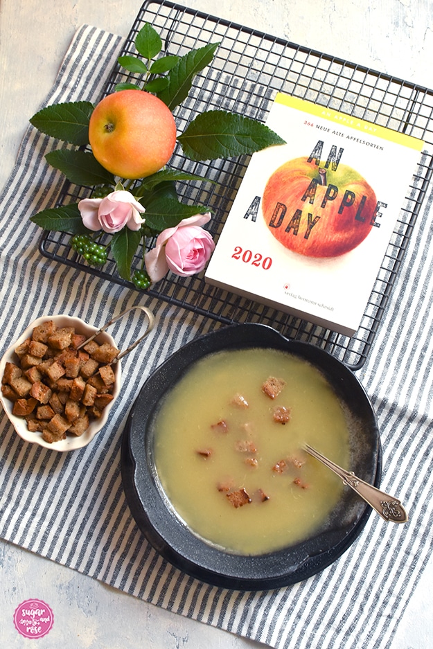 Graue Steingutschale mit Apfelkoch, darin ein alter Löffel, daneben eine kleine weiße Schale mit gerösteten Brotwürfeln und einer Brotzange, dahinter ein schwarzes Brotgitter, darauf ein Apfel und zwei kleine rosa Röschen. Am Gitter liegt der neue Tagesabreißkalender "An Apple a day 2020", auf dem in der Mitte ein rotgelb gestreifter Apfel abgebildet ist, darüber der Kalendertitel in gebrochenen schwarzen Lettern