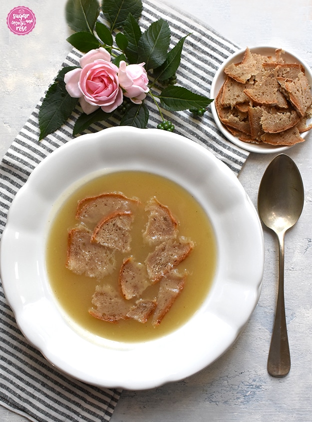 Weißer, alter Steinguteller mit Apfelkoch, daneben ein großer alter Suppenlöffel, dahinter ein kleines weißes Metallschälchen mit dünn aufgeschnittenen Brotschnitten, alles auf einer grau-weiß gestreiften Serviette mit rosa Rosenblüten und grünen Blättern dekoriert