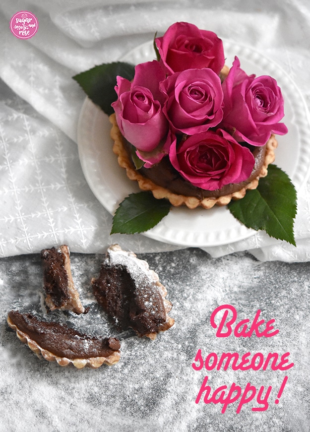 Ein Schokotartelette auf weißem Vintageteller mit fünf pinkfarbenen Rosen in Großaufnahme auf weißem Stofftuch, dieses auf mit Puderzucker dicht bestreuter Arbeitsplatte und ein angebrochenes Stück eines Tartelettes. Auf dem Bild steht rechts unten in pinkfarbener Schrift "Bake someone happy!"