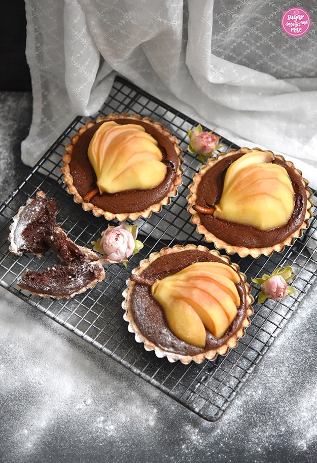 Drei kleine Metallförmchen mit Birnen-Schoko-Tartelettes auf einem Gitter, mit Puderzucker bestreut und kleinen rosa Röschen dekoriert.