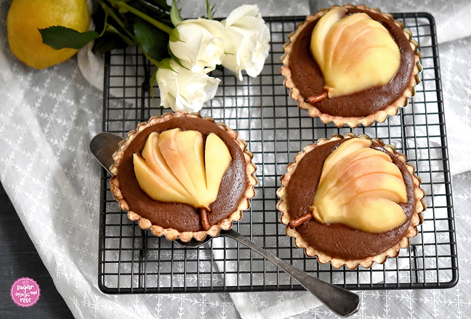 Drei Birnen-Schoko-Tartelettes auf einem schwarzen quadratischen Gitter, welches auf einem weißen Stoffband steht, daneben drei weiße Rosen und eine gelbe Birne