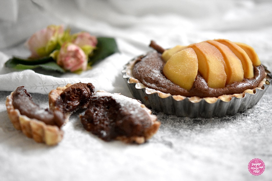 Frontalansicht eines Birnen-Schoko-Tartelettes, vorne ein angebrochenes Tartelette, wo man den weichen Schokokern sieht, rundum reichlich Puderzucker