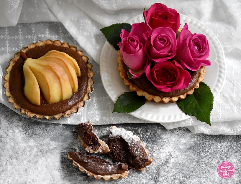Ein Birnen-Schoko-Tartelette, daneben eine Tartelette auf weißem Vintage-Dessertteller, dekoriert mit fünf pinkfarbenen Rosenköpfen, davor ein Stück von einem Schokotartelette