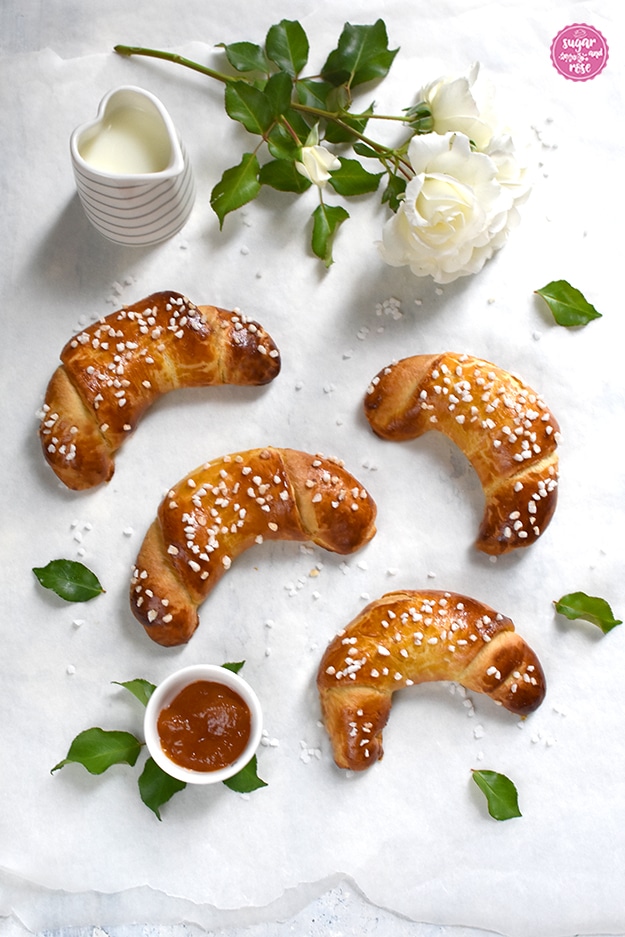 Vier Briochekipferl auf weißer Tischplatte mit einer weißen Rose mit mehreren Blüten, daneben ein herzförmiges Milchkännchen und eine kleine Schale mit Marillenmarmelade im Vordergrund