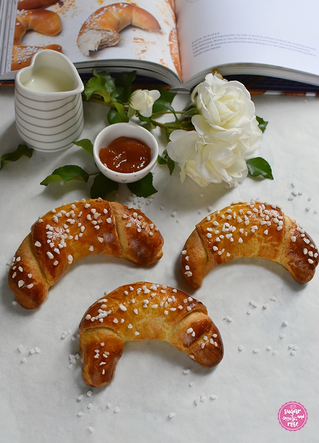 Aufgeschlagenes Kochbuch mit abgebildeten Kipferln, davor eine weiße Rosenblüte, ein herzförmiges Milchkännchen, ein Marmeladeschälchen und drei Briochekipferl auf weißem Untergrund, der mit Hagelzucker bestreut ist.