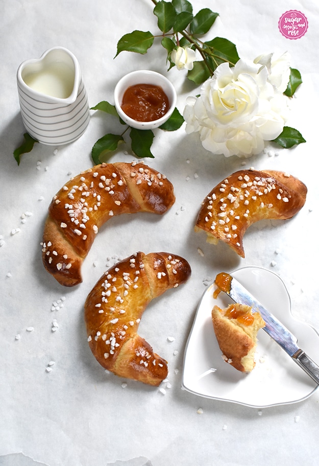 Zwei ganze und ein angebrochenes Briochekipferl auf weißer Tischplatte mit einer weißen Rose, daneben ein herzförmiges Milchkännchen und eine kleine Schale mit Marillenmarmelade sowie ein herzförmiger Teller mit einem Stück Brioche, ein Messer mit etwas Marmelade