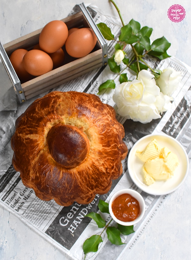 Auf einem alten Zeitungspapier liegend, von oben fotografiert, eine große Kopfbrioche, eine kleine Holzkiste mit Metallgriffen mit 6 braunen Eiern, ein Butterteller, ein Marmeladeschälchen und weiße Rosen