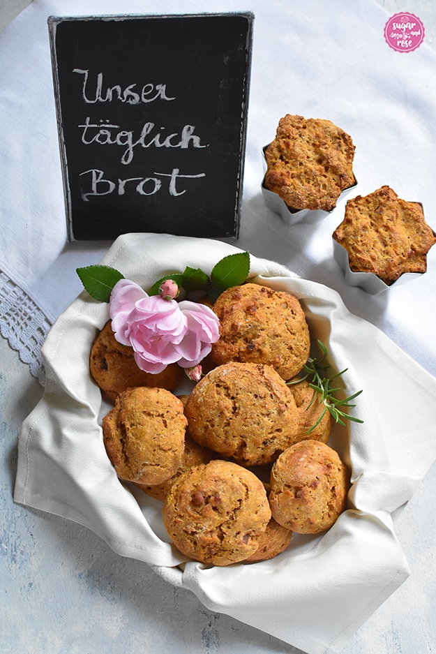 Ein Brotkörbchen mit hellem Tuch, darin mehrer leicht orangefarbene Kürbis-Speckweckerl und eine rosas Rosenblüte, dahinter ein kleine Tafel, beschriftet mit Kreide: "Unser täglich Brot"