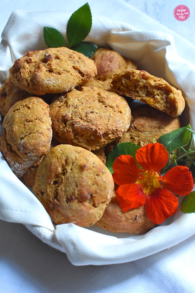 Kürbis-Speckweckerl im Brotkörbchen in Großaufnahme, ein angebrochenes Brötchen und eine orangefarbene Kapuzinerkresseblüte mit grünen Blättern