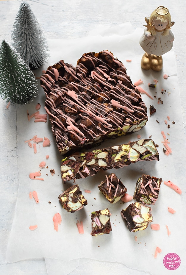 Dunkles Rocky Road auf weißem Backpapier, garniert mit rosa Schokoresten, dahinter eine stehende Engelfigur und zwei grüne Dekotannenbäumchen