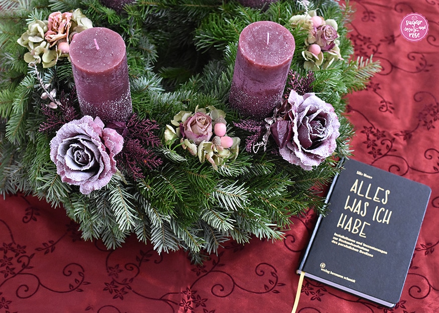 Adventskranz auf weinrotem Damasttischtuch, davor ein schwarzes Buch mit Goldlettern "Alles was ich habe"