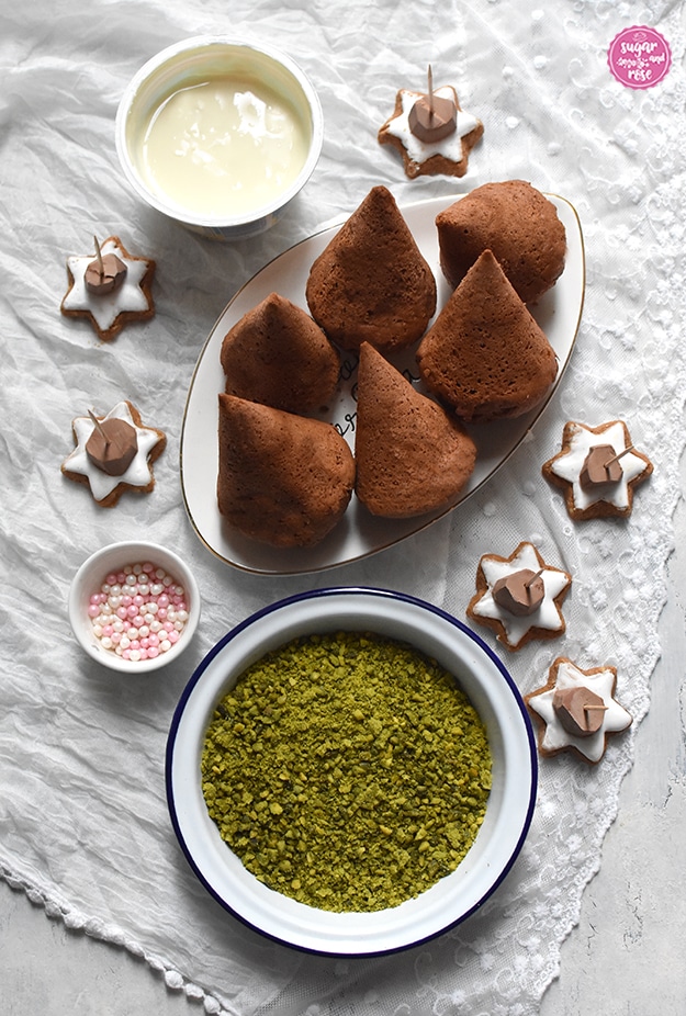 Fertig gebackene Lebkuchenbäumchen auf Teller, davor eine Schüssel mit gehackten Pistazien, dahinter ein Becher flüssige weiße Kuvertüre, ein kleines Schüsselchen mit rosa und weißen Zuckerperlen, rundum Zimtsterne mit Zahnstochern
