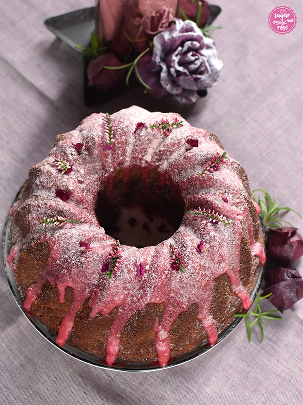 Punschgugelhupf mit rosa Zuckerguss auf Leinenserviette, dahinter eine rosegoldene Kerze auf einem Sternenteller, dekoriert mit einer auberginefarbenen Kunstrose mit Schneestaub