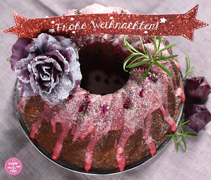 Punschgugelhupf von oben, dekoriert mit einer auberginefarbenen Seidenblüte mit Schneestaub, Rosmarinzweigen und getrockneten dunkelrosa Rosenblüten, on the top ein weinrotes Schildchen mit der Aufschrift "Frohe Weihnachten"