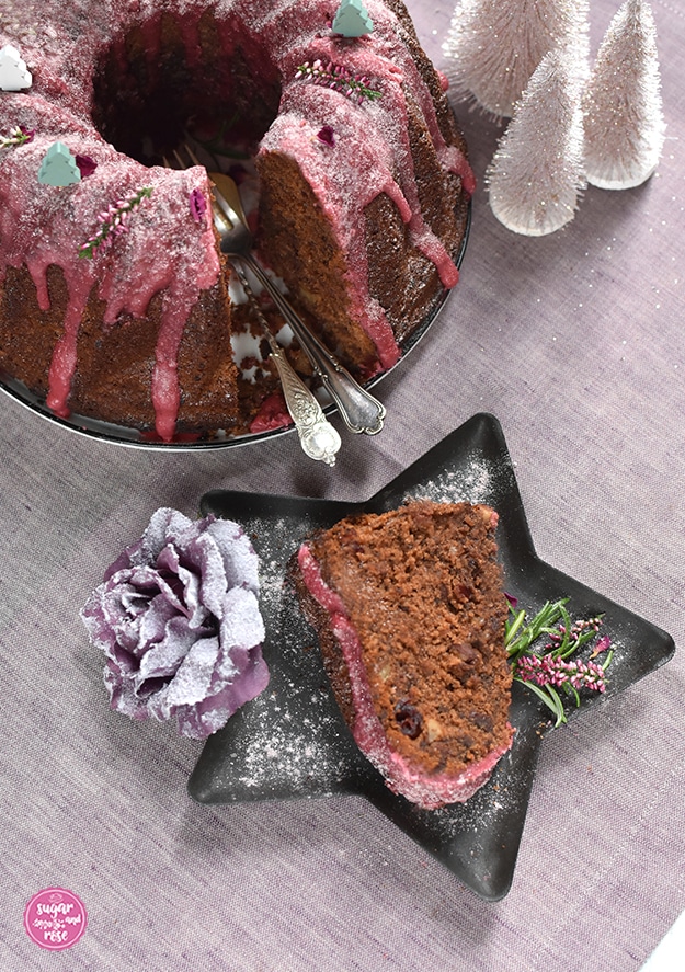 Ein Stück Punschgugelhupf mit Schokostückchen, Nüssen und Cranberryeinschlüssen auf ein einem Sternenteller, daneben Rosmarin und Erikablüten, dahinter der angeschnittene Gugelhupf mit Gabel und Löffel am Teller liegend